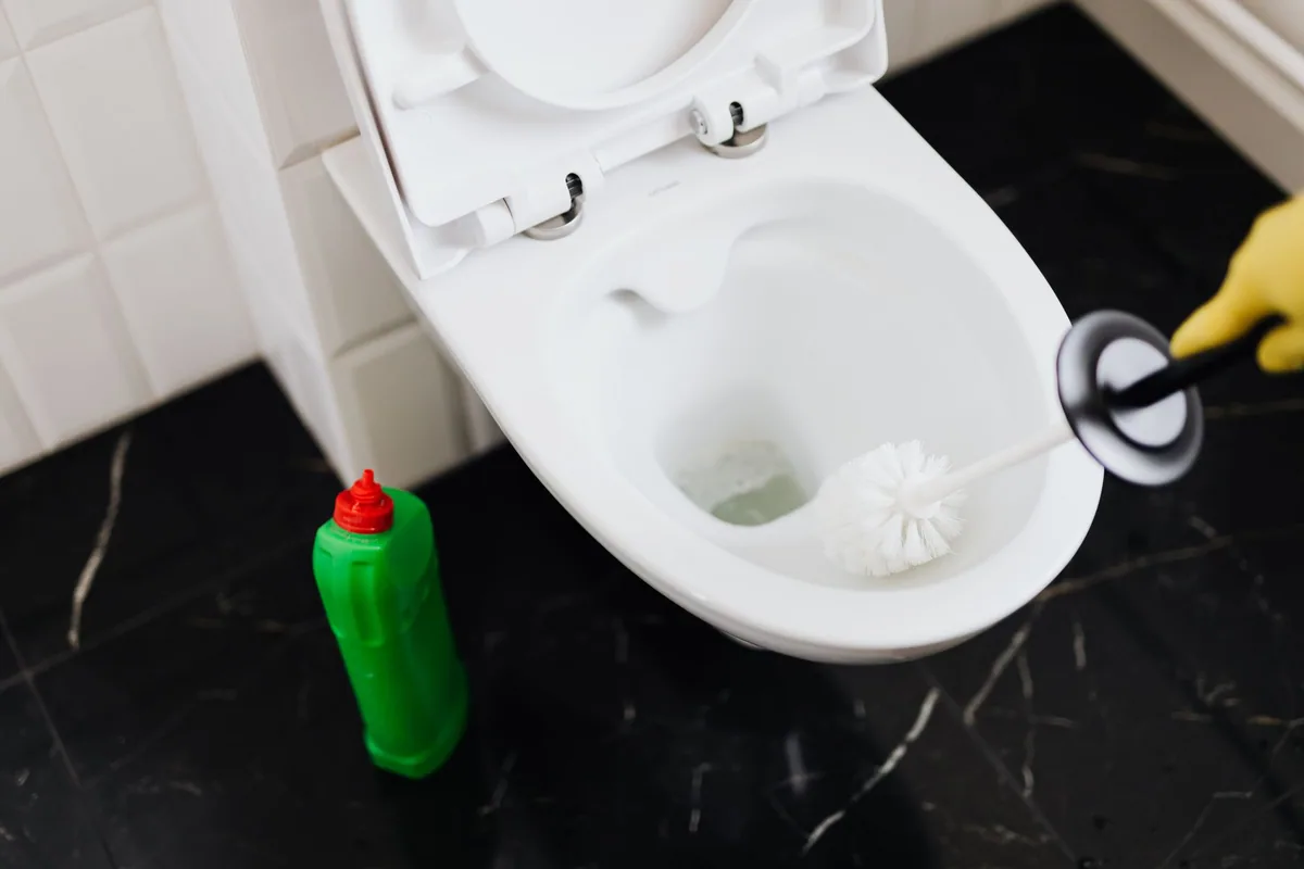 WC con la tapa levantada, una escobilla y una botella de limpiador en el suelo del baño
